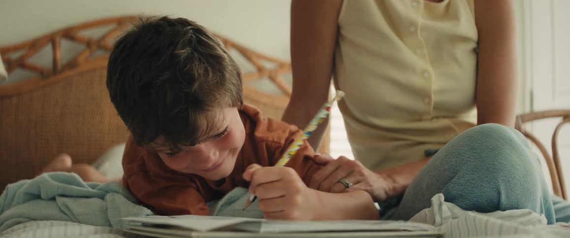 Close-up of a smiling boy drawing in a notebook on his bed as his mother affectionately kisses his head