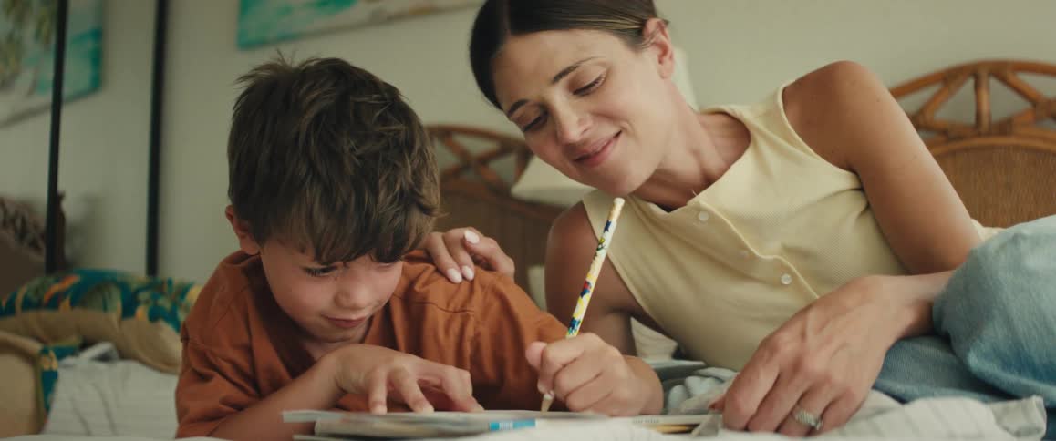 A proud mother helps her young son with his homework as they laugh and smile together while lying on a bed