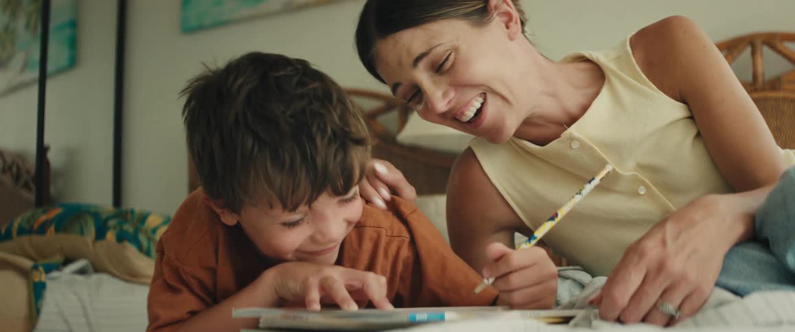 A proud mother helps her young son with his homework as they laugh and smile together while lying on a bed