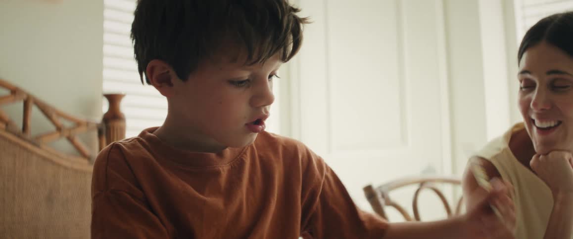 A proud mother lovingly watches her young son as he concentrates on drawing at home