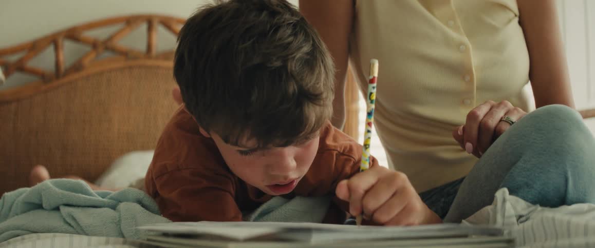 A young boy carefully writes in a notebook while his mother sits beside him on a bed and helps him learn