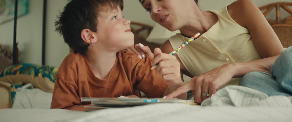 A smiling mother helps her young son draw in a coloring book while they relax on a bed in a brightly lit room