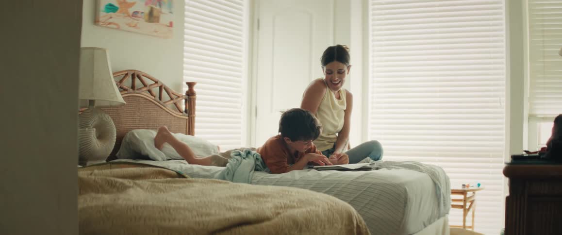 A smiling mother watches her young son draw in a book on a bed, sharing a high-five and a loving kiss in a sunlit room