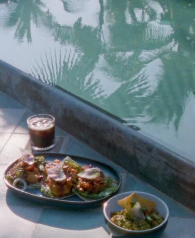 A vintage-style shot of delicious tacos and guacamole served with a cold drink by a tranquil swimming pool reflecting palm trees