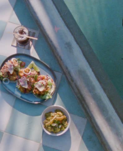 A high-angle, vintage-style shot of delicious tacos, guacamole, and a drink sitting next to a sunlit swimming pool