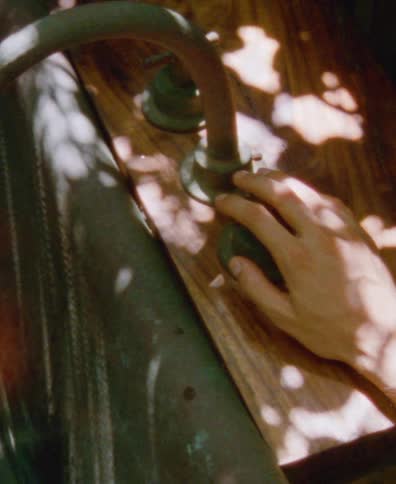 A hand turns the handle of a vintage outdoor faucet, causing water to flow into a copper basin in dappled sunlight