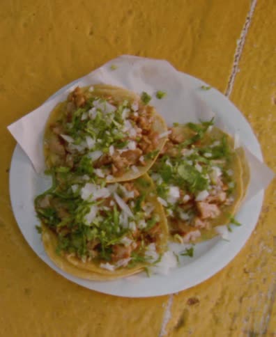 A shaky, close-up, top-down view of three traditional Mexican street tacos served on a white plate against a bright yellow background
