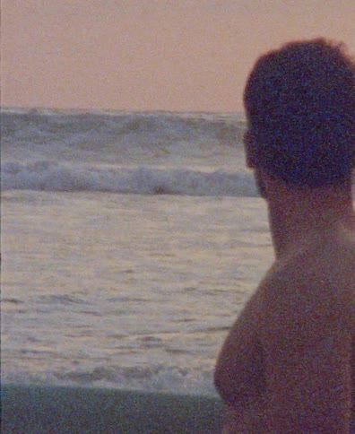 A grainy, vintage-style film captures a man from behind as he stands on the beach watching the ocean waves crash during a beautiful pink sunset