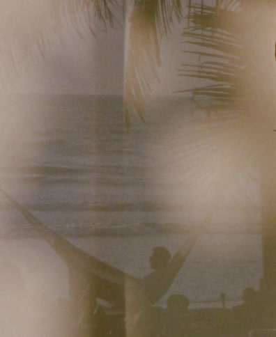 A nostalgic, sepia-toned clip of a person relaxing in a hammock under palm trees while looking out at the ocean