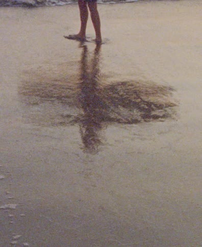 The reflection of a surfer standing in the shallow ocean water is distorted by gentle incoming waves on the sandy shore