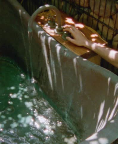 Dappled sunlight dances on an outdoor copper bathtub as a person's hand rests by the running faucet