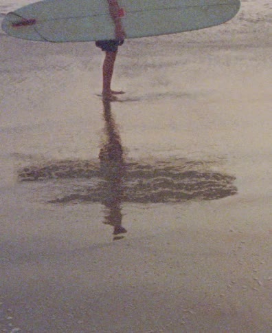 The distorted reflection of a surfer holding their board creates a cross shape on the wet sand in this grainy, vintage-style clip