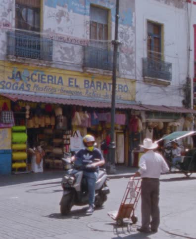 Pedestrians, a taxi, and scooters navigate a bustling street corner in Mexico in a vintage, grainy film shot