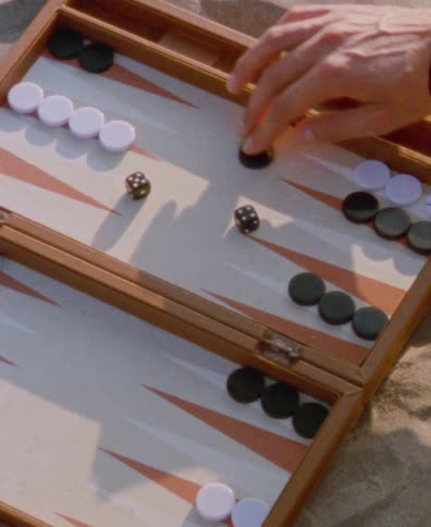 In a slow-motion close-up, a person's hand rolls dice and moves pieces across a backgammon board resting on the sand