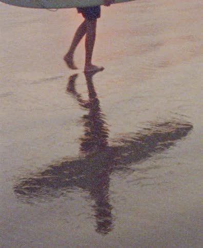 A surfer's reflection distorts in the shallow waves as they walk barefoot along the sandy shore with their surfboard at sunset