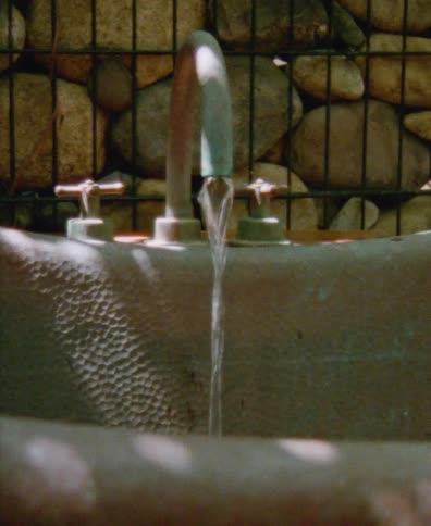 A hand turns on the faucet of a rustic hammered metal sink, letting the water flow in a sun-dappled outdoor setting