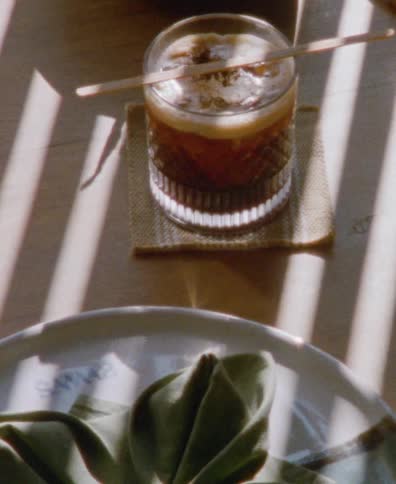 An iced coffee sits on a table in the morning sun, with light and shadows from blinds creating a striped pattern