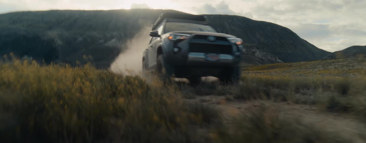 A rugged black SUV with a roof tent drives off-road on a dusty trail through a grassy, mountainous landscape at golden hour