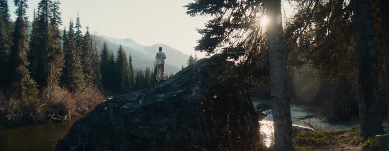 A nature photographer hikes to the top of a large boulder next to a river to capture photos of the scenic mountain landscape at sunrise