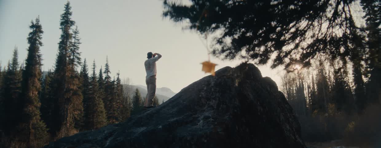 A photographer hikes up a large rock in the wilderness to capture the mountain landscape with his camera at golden hour