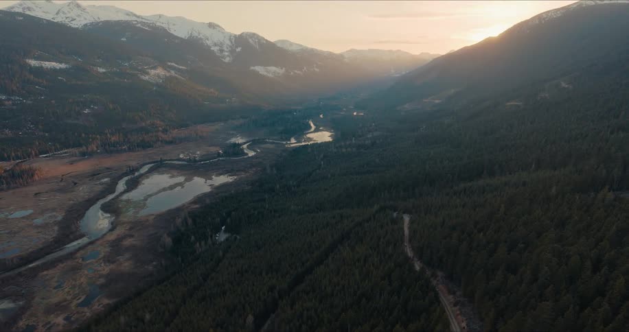 A static aerial shot captures the majestic beauty of a vast mountain valley with dense forests and a winding river at sunset