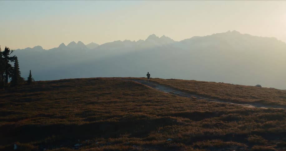 A trail runner jogs along a scenic mountain path toward a distant, snow-capped mountain range during a golden sunset