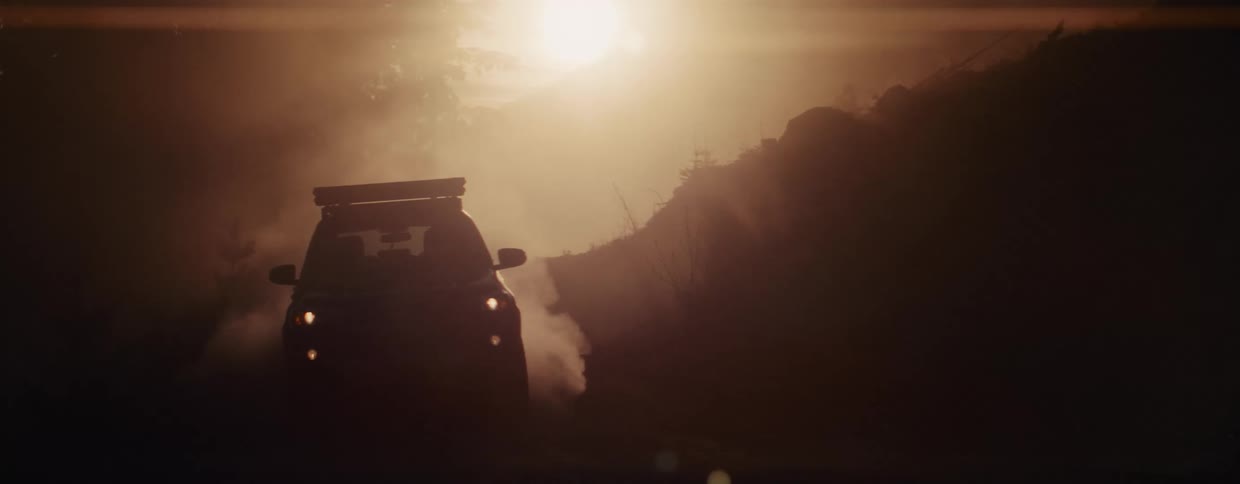 An SUV with a rooftop tent drives towards the camera on a dusty road at sunset, kicking up a cloud of smoke