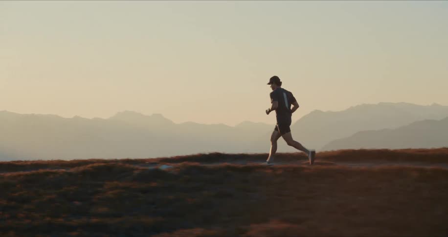 A male athlete runs along a high mountain trail in slow motion, with a stunning mountain range backdrop at golden hour sunset