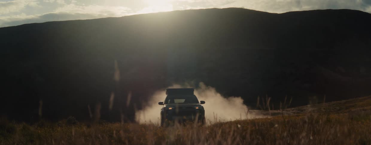 An overlanding SUV drives through a dusty mountain pass at golden hour, kicking up a large cloud of dust in slow motion