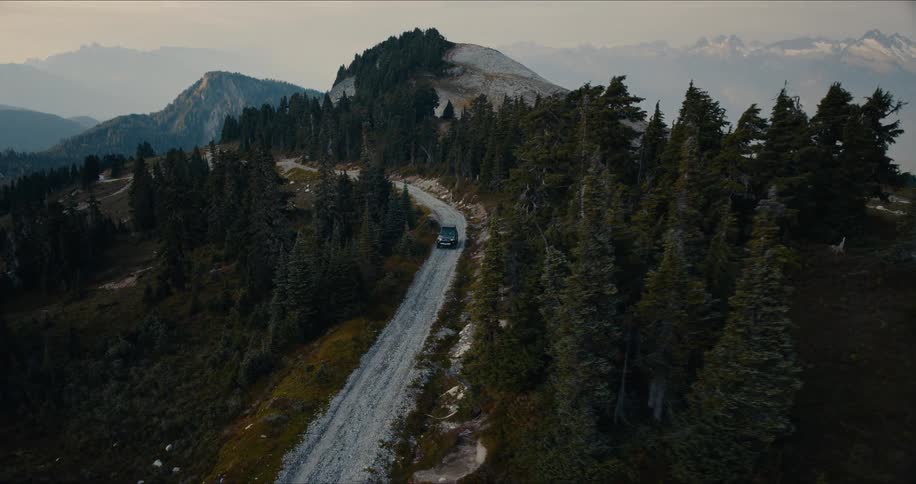 An aerial view shows an SUV driving along a remote gravel road on a mountain ridge surrounded by a vast, tree-covered landscape