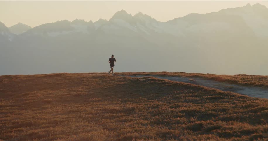 A male trail runner jogs in slow motion along a mountain ridge during a beautiful golden sunrise with a vast, snow-capped mountain range in the background
