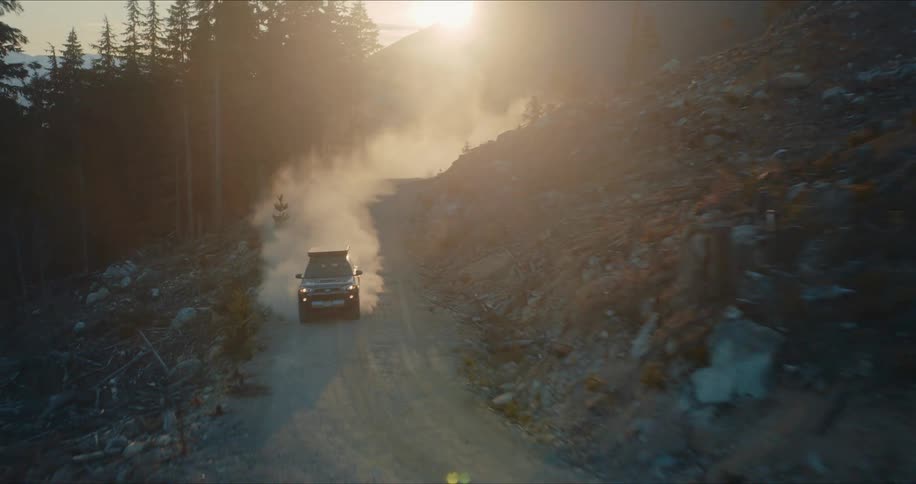 An aerial shot follows an SUV driving on a dusty, remote mountain road, kicking up a dramatic plume of dust in the golden hour light