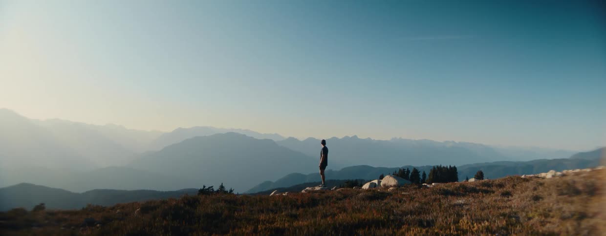 A lone hiker with a backpack stands on a mountain peak, turning on his headlamp as he overlooks a vast, hazy mountain range at dawn