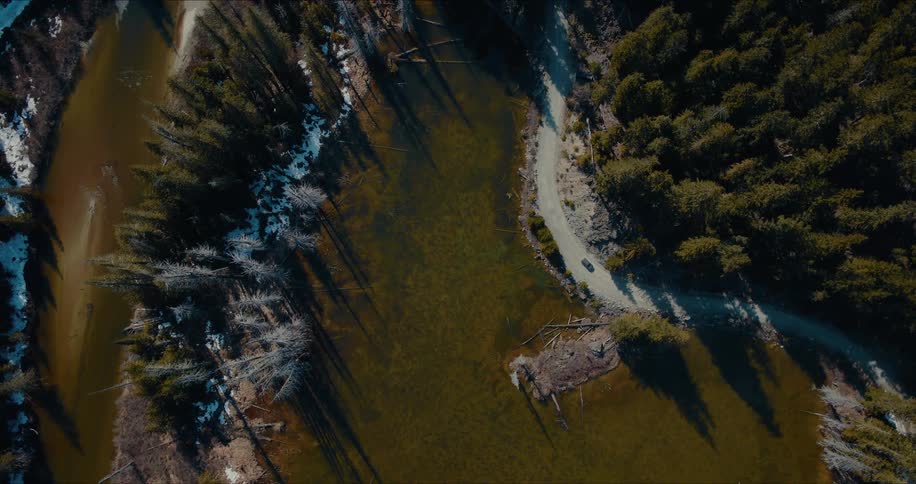 Aerial top-down view of a car driving on a remote dirt road alongside a lake through a forest in early spring