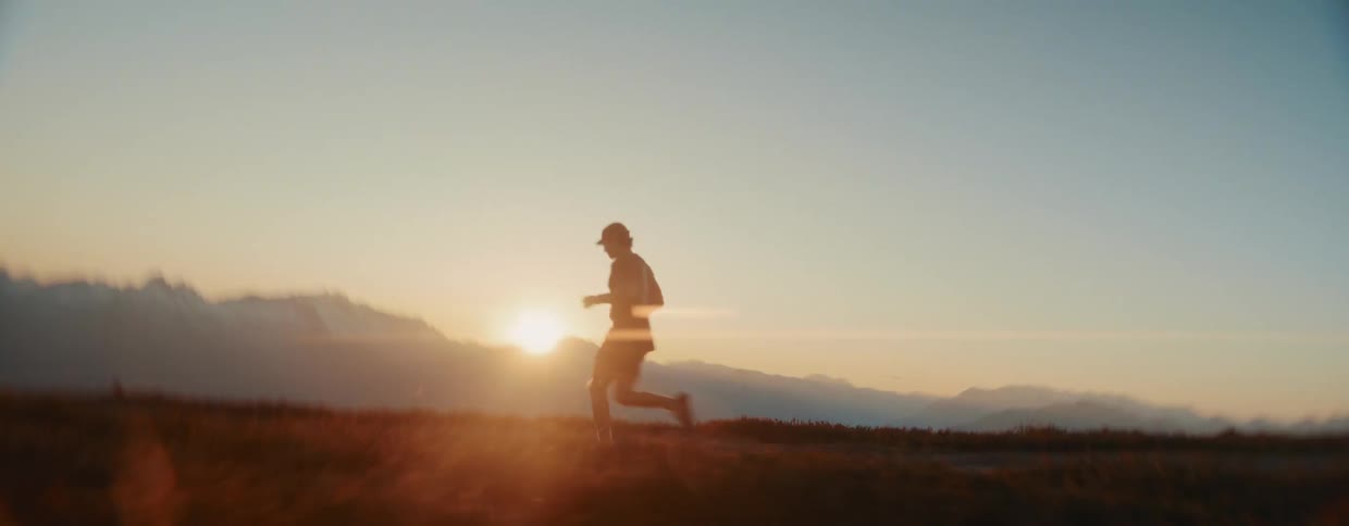 An athletic young man trail runs along a scenic mountain ridge, silhouetted against the beautiful golden hour sunset