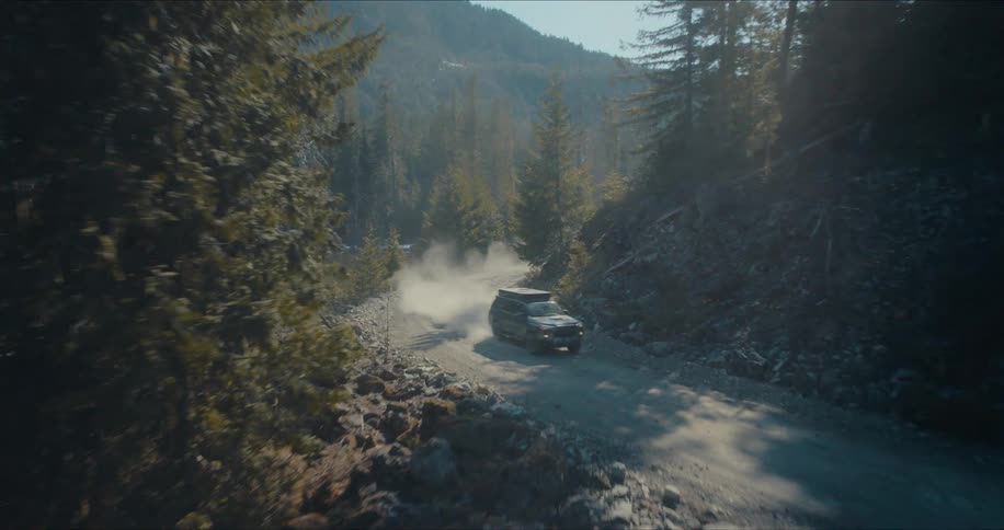 An aerial shot follows a rugged SUV as it kicks up dust while driving on a winding dirt road through a mountain forest next to a lake