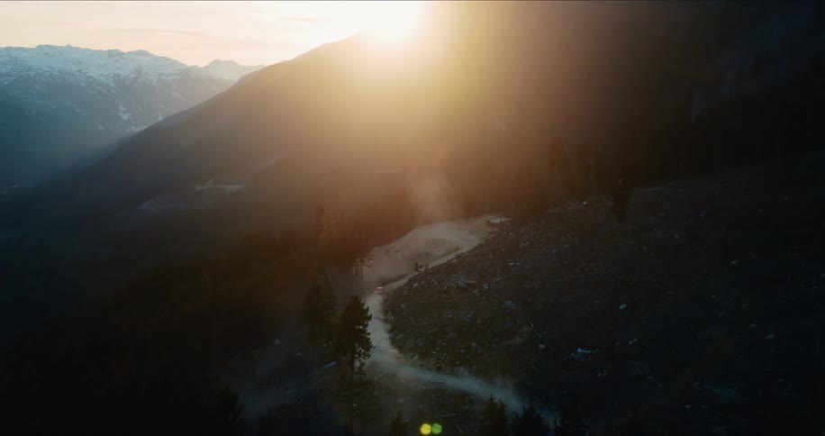 Epic aerial view of a car driving on a winding dirt road through the mountains as the sun rises over the peaks