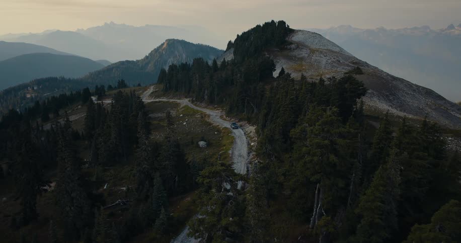 A lone car navigates a winding dirt road through a vast, tree-covered mountain range in this stunning aerial shot at sunset