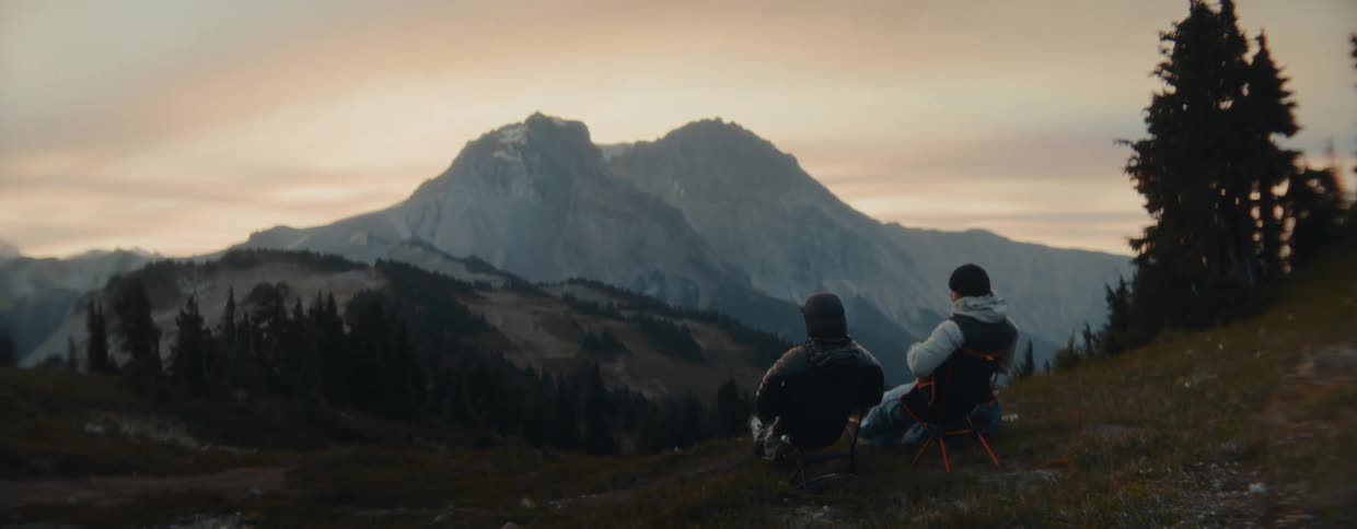 Two male friends relax in camping chairs on a scenic overlook, talking and pointing at the majestic mountains during a beautiful sunset