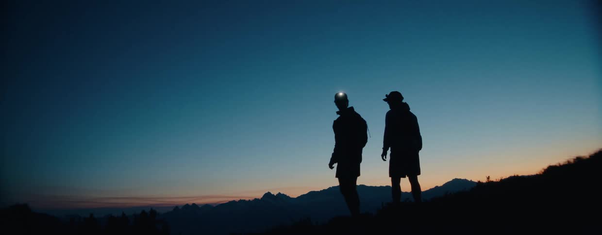 Two silhouetted hikers celebrate reaching the summit with a fist bump and an embrace as they admire the mountain vista at twilight