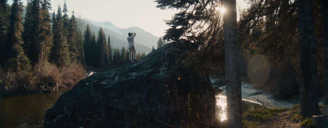 A lone photographer climbs onto a large boulder next to a river to capture the scenic mountain landscape at sunrise