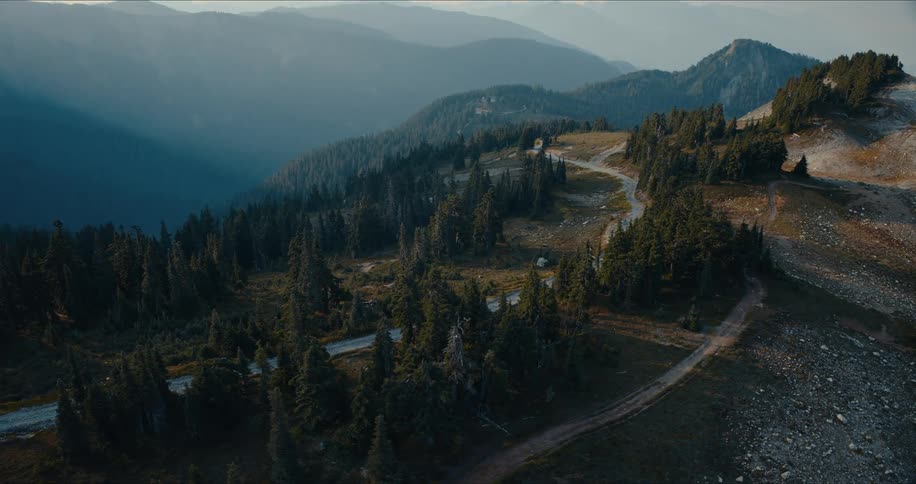 An epic aerial shot follows a lone SUV driving along a winding dirt road on a vast, tree-covered mountain pass at sunrise