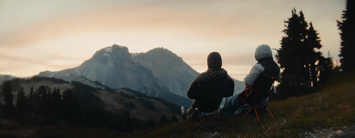 Two adventurous campers sit in chairs on a scenic mountain overlook, pointing towards the majestic range as the sun rises