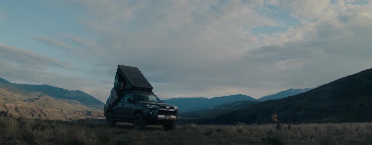 A couple camps with their SUV and rooftop tent, enjoying the serene, wide-open mountain scenery at dusk
