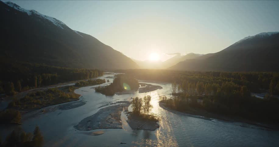 A breathtaking aerial shot flies over a dense forest to reveal a majestic river valley between snow-capped mountains at sunrise
