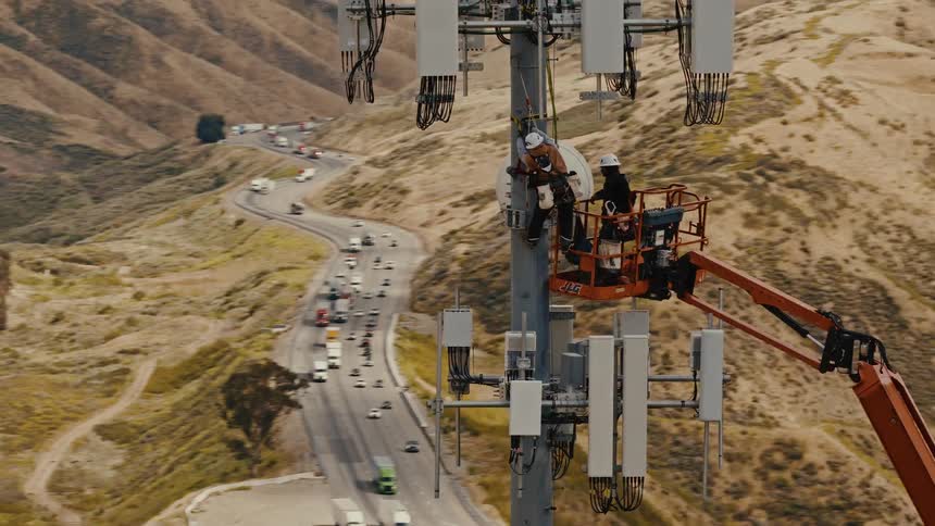 Aerial view of two technicians in a boom lift working on a cellular tower high above a busy freeway in the hills
