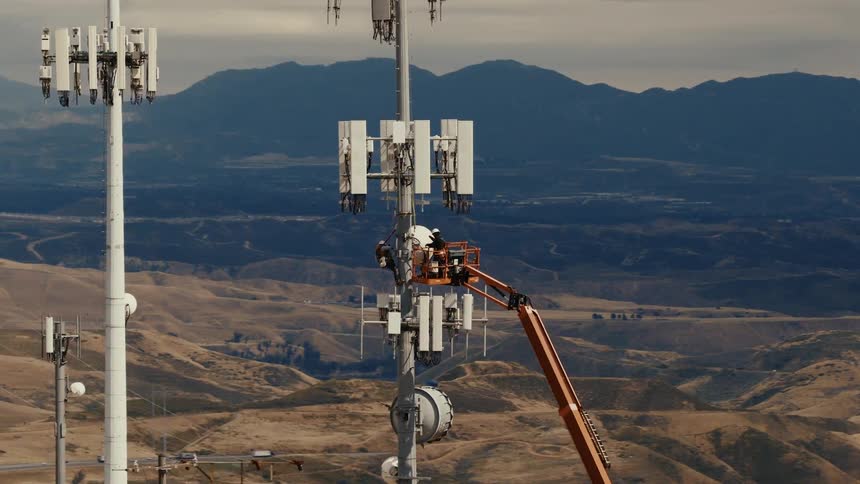 Aerial view of technicians on a cherry picker performing maintenance on a cellular communications tower in a mountainous region
