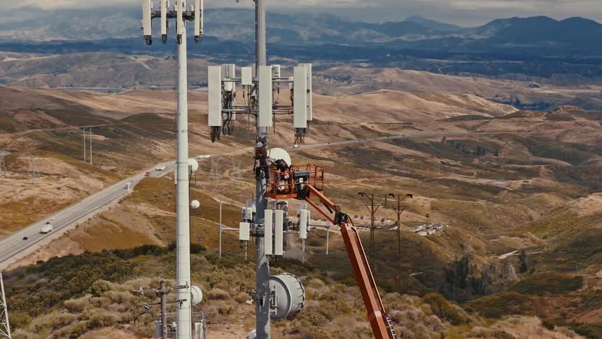 Aerial view of workers in a cherry picker performing maintenance on a telecommunications tower with a scenic mountain pass in the background