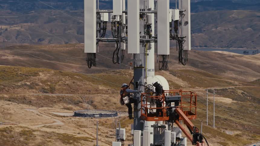 An aerial view captures technicians carefully performing maintenance on a tall 5G cell tower amidst a vast, hilly landscape