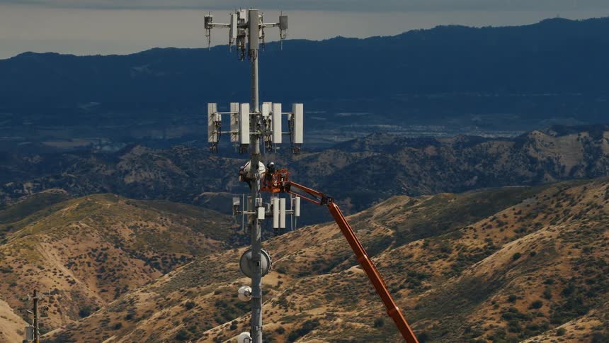 Telecommunication workers service a cell tower from a crane high on a mountain with a vast landscape below